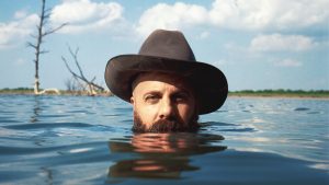 Marty Bush in a body of water with only the top half of his head above water, wearing a grey cowboy hat.