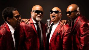 Members of the Blind Boys of Alabama pose in red suit jackets around a microphone.