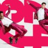 Two dancers from Chicago Tap Theatre bend and leap. They are wearing pink suits with white dress shirts and are in front of the numbers 23-24.