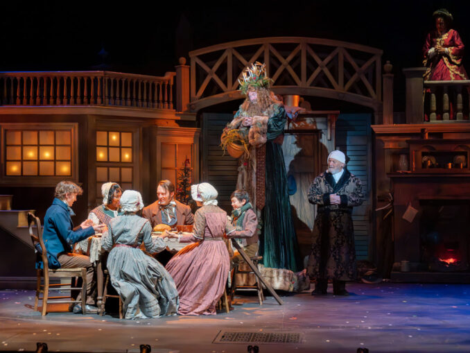 The Cratchit family enjoys Christmas dinner, while Scrooge and the Ghost of Christmas Present look on. • The cast of KCRep's 2023 production of A CHRISTMAS CAROL • Photo by Don Ipock