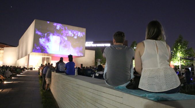 In the foreground two people attend a concert on the lawn of Nerman Museum. Images are displayed on the exterior wall of the museum.